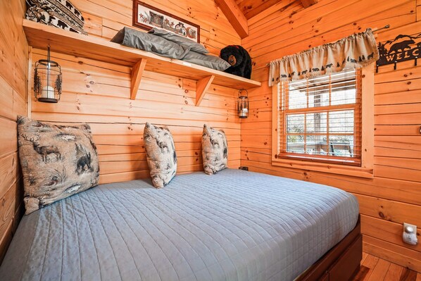 A wooden cabin room with a bed, a shelf with a bear plush toy, and a window with a 'WELCOME' sign. - Snuggle Bear | Hocking Hills, Adorable Tiny Home (Logan)