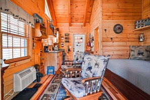 A log cabin interior with a wooden floor, a sofa, a bed, a kitchen area, and a window with blinds. - Snuggle Bear | Hocking Hills, Adorable Tiny Home (Logan)