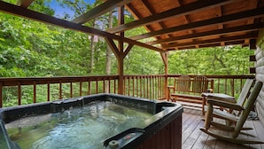 A wooden deck with a hot tub, wooden chairs, and a view of a forest.