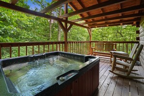 Un jacuzzi en una terraza de madera con una mecedora y una mesa, rodeado de abundante follaje.
