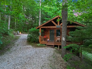 A wooden cabin with a porch, surrounded by trees and a gravel path.