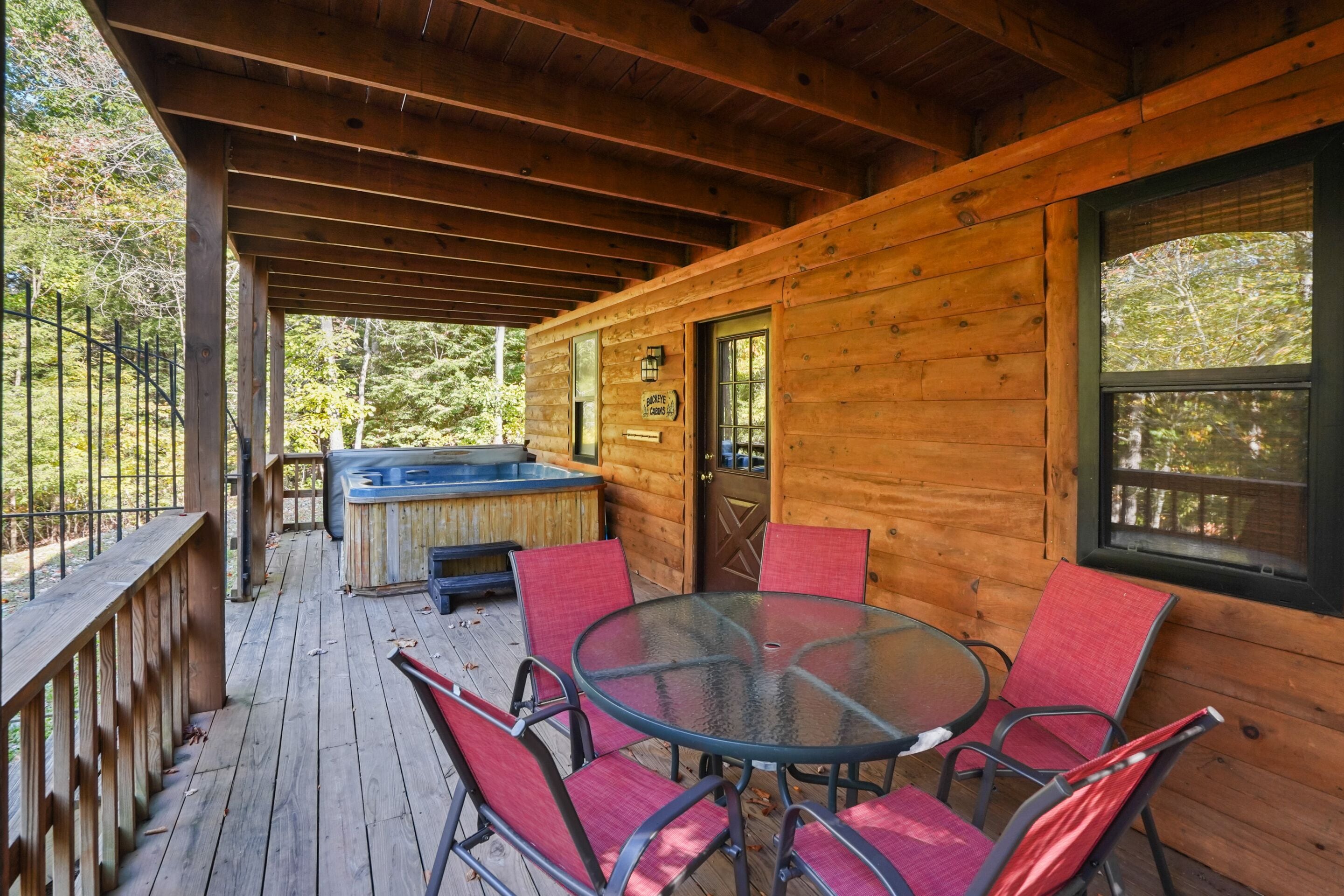 A wooden cabin with a glass table and red chairs, a hot tub, and a deck with a railing.