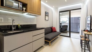 A modern kitchen with a built-in microwave, a sink, and wooden cabinets. There is a gray sofa with a red pillow and a small table with stools. A sliding glass door leads to a balcony. -  Charlie Alves Guimarães Pinheiros (São Paulo)