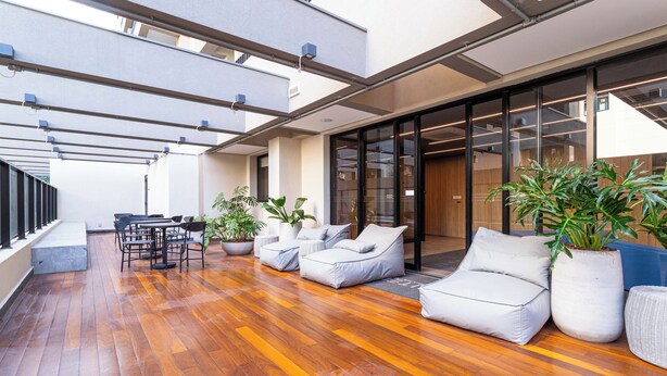 A modern outdoor seating area with wooden flooring, white cushioned sofas, and potted plants. -  Charlie Alves Guimarães Pinheiros (São Paulo)