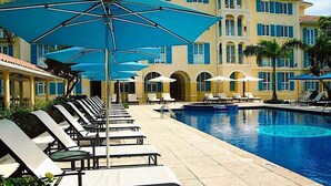 Un espace aménagé au bord de la piscine, avec des chaises longues et des parasols, une piscine, et un bâtiment à la façade jaune et aux volets bleus.