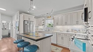 A modern kitchen with white cabinetry, a central island with stools, stainless steel appliances, and a backsplash with a mosaic tile design. - Kiss Me Kottage- Private Waterfront Home! (Cedar Key)
