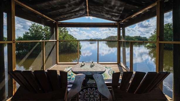 A covered outdoor seating area with a view of a calm lake and trees. - Kiss Me Kottage- Private Waterfront Home! (Cedar Key)