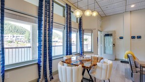 A dining area with a wooden table and white chairs, large windows with blue curtains, and a view of a building and trees. - Keystone Lake Haven: Heated Pool & Ski Shuttle! (Keystone)