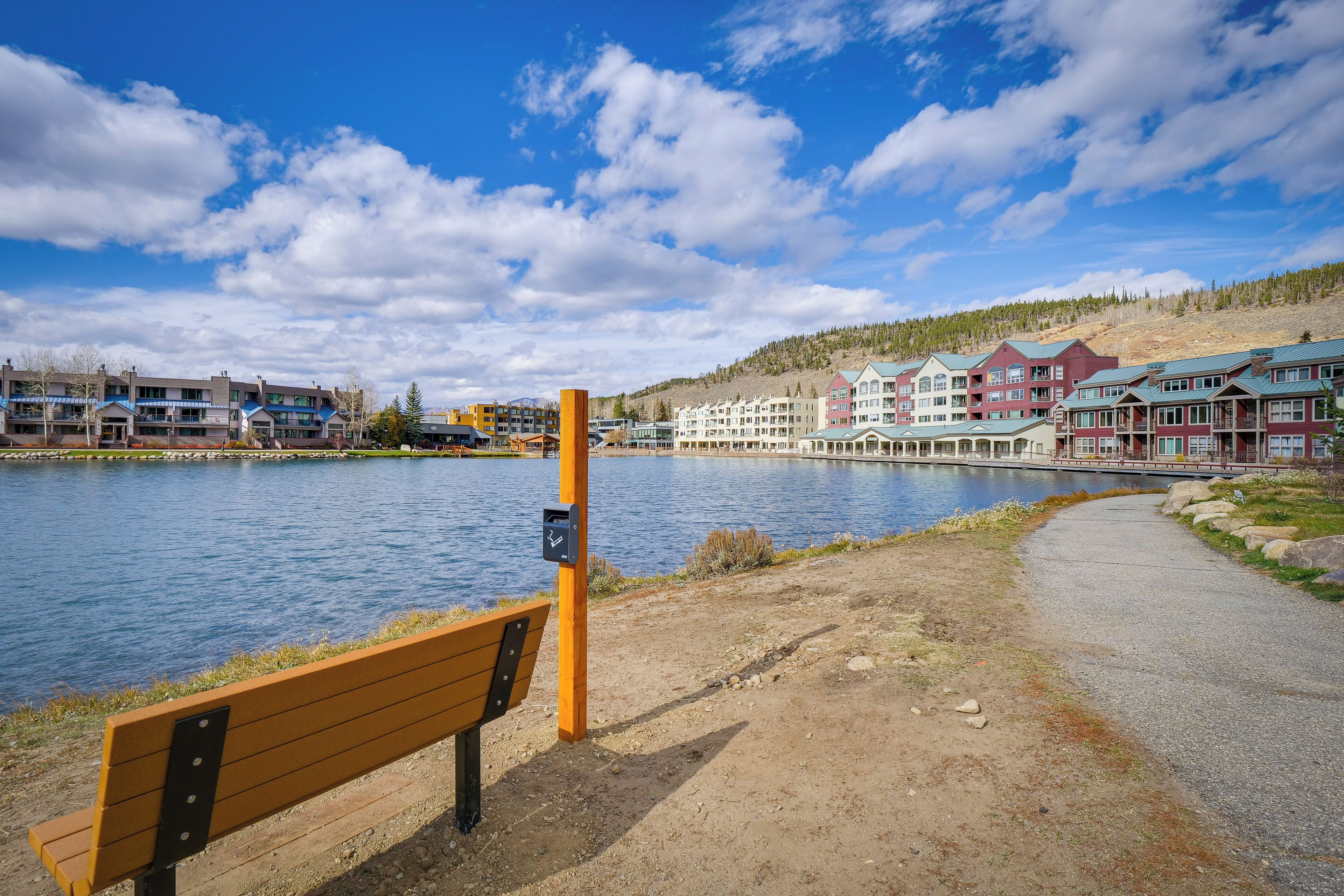 A lakeside view with a wooden bench, a path, and buildings in the background.