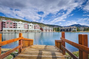En träbrygga som leder fram till ett hotell vid vattnet, bestående av flera byggnader, med en lugn sjö och berg i bakgrunden.