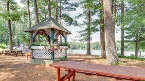 En pergola med gittertop og blomsterdekorationer, omgivet af picnicborde og bænke, beliggende i et skovområde med en sø i baggrunden.
