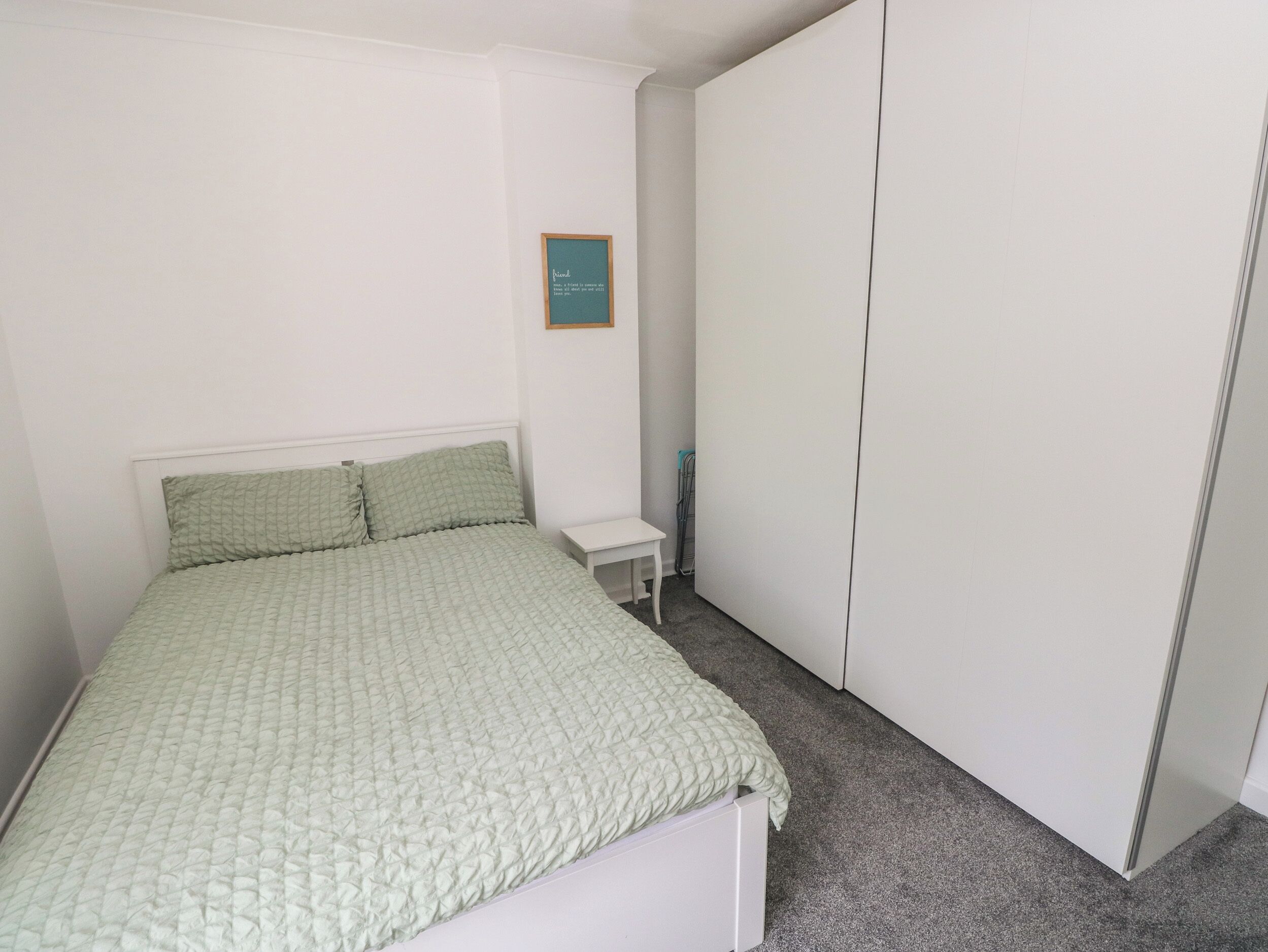 Un dormitorio con cama, una mesita de noche pequeña, un armario y una cita enmarcada en la pared.