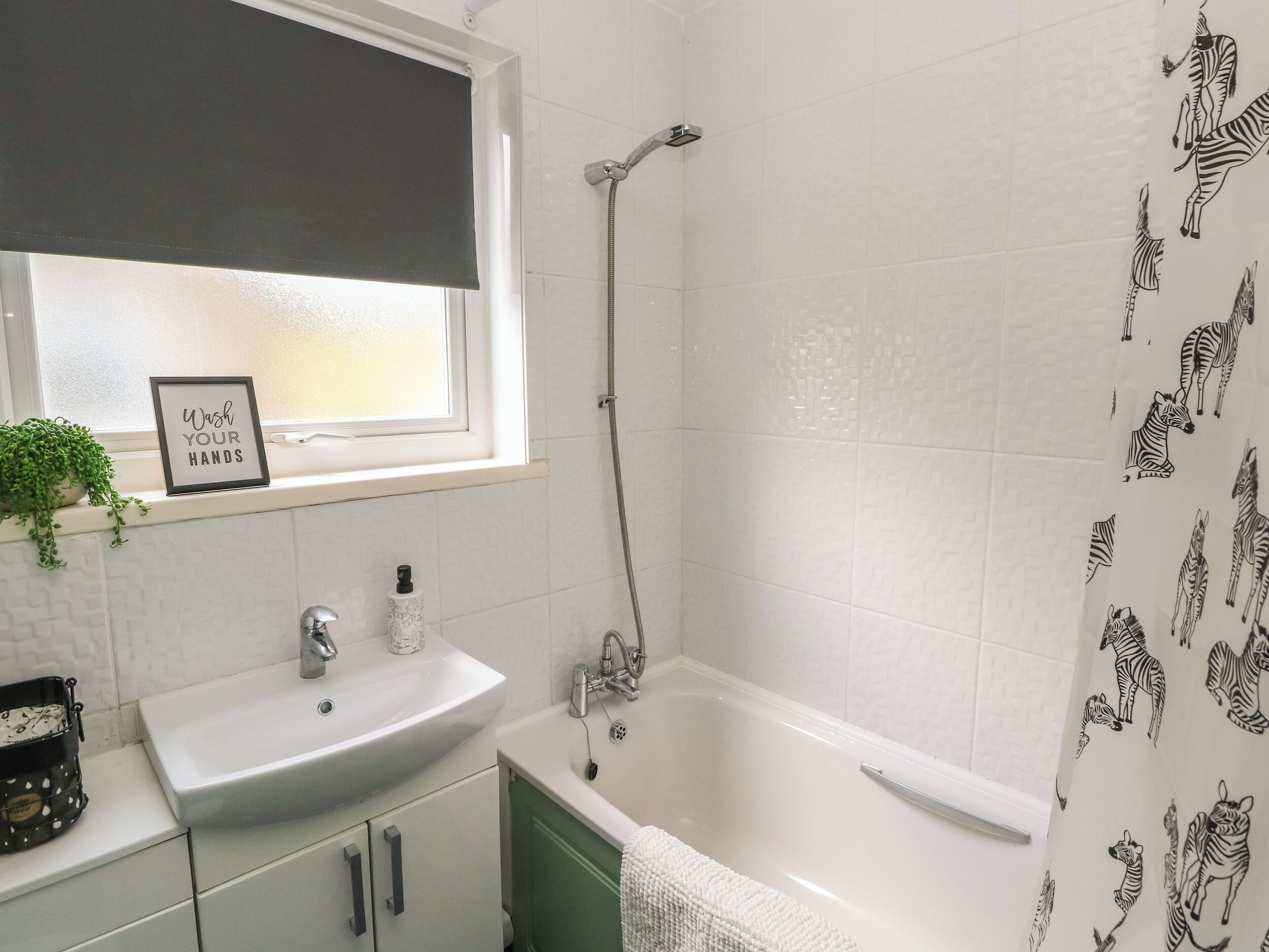 A bathroom with a white sink, green cabinet, and a bathtub. There is a window with a black blind and a sign that says "Wash Your Hands".