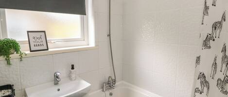 A bathroom with a white sink, green cabinet, and a bathtub. There is a window with a black blind and a sign that says "Wash Your Hands".