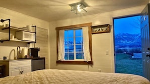 Une chambre avec un lit, une fenêtre avec des rideaux, un four à micro-ondes posé sur une étagère, un évier et une porte donnant sur un balcon avec vue sur les montagnes.