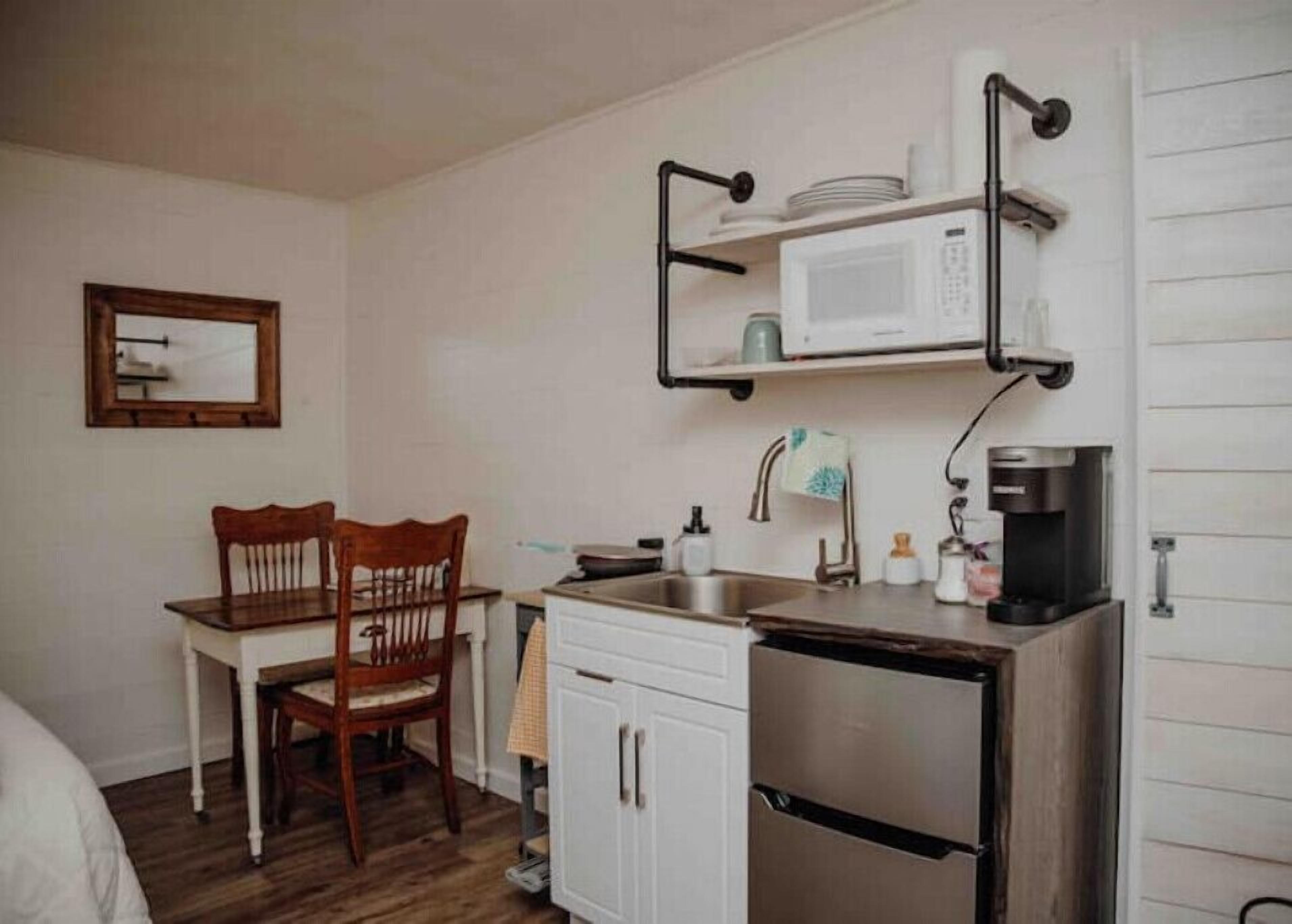 A compact kitchenette with a microwave, coffee maker, and a small sink. There is a wooden chair and a white cabinet.