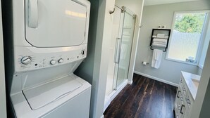 A laundry area with a washer and dryer, a glass shower enclosure, and a window with a view of trees.