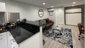 Ein modernes Wohnzimmer mit einer Kücheninsel aus schwarzem Marmor, einem grauen Sofa mit rotem Kissen und einem blau-weißen Teppich.