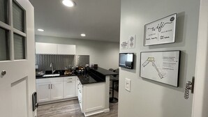Eine moderne Küche mit weißen Schränken, einer schwarzen Arbeitsplatte und einer Spritzschutzwand. An der Wand befinden sich zwei wandmontierte Fernseher und ein gerahmter Notfallplan.