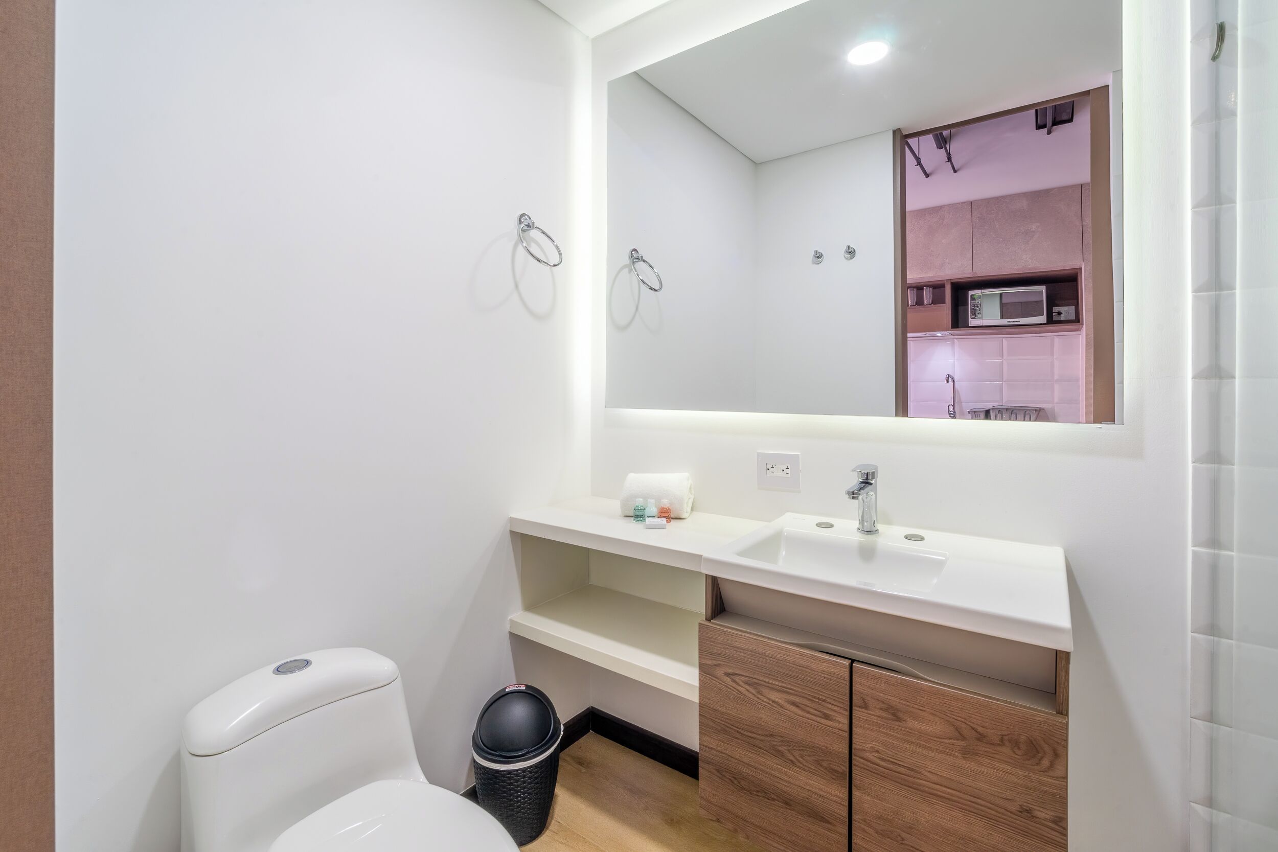 Un baño moderno con un lavamanos blanco, un mueble de madera y un espejo que refleja una zona de cocina americana.