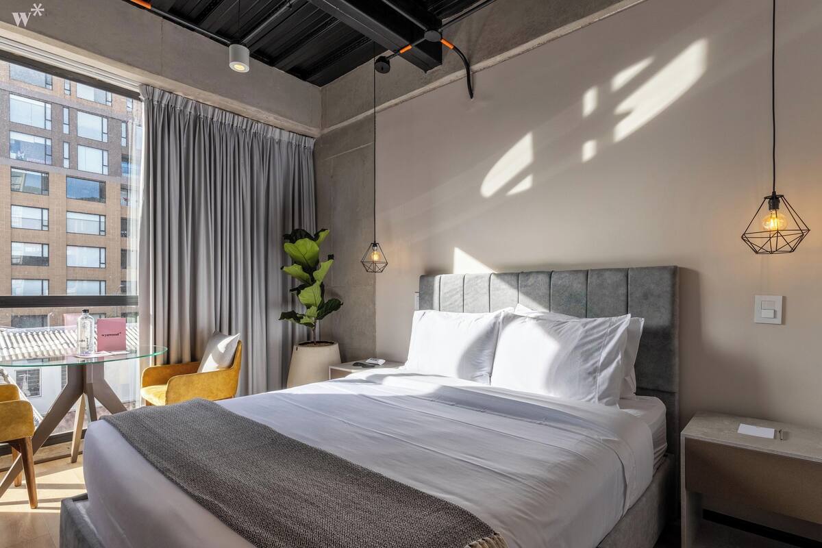 Habitación de hotel moderna con una cama grande, una mesa de vidrio, una silla, una planta en maceta y vistas a la ciudad.