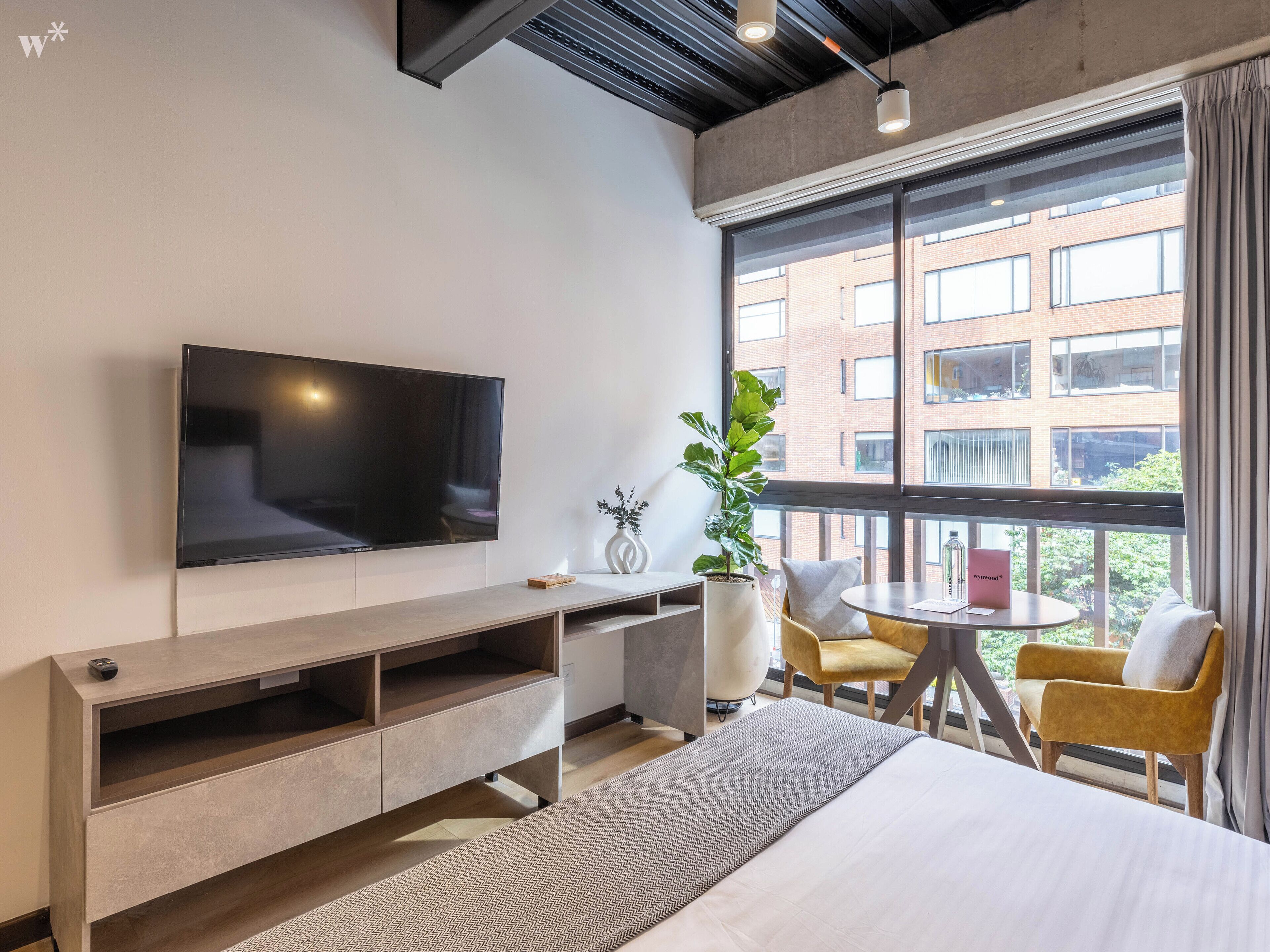 Ein modernes Hotelzimmer mit einem großen Flachbildfernseher, einem Bett, einem kleinen runden Tisch mit Stühlen, einer Topfpflanze und Blick auf ein Backsteingebäude durch große Fenster.