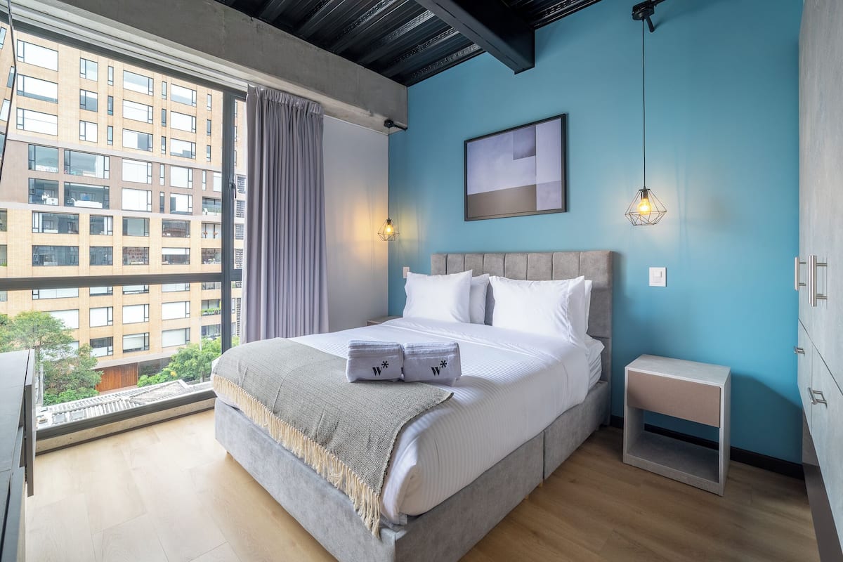 Un dormitorio moderno con una cama grande, vista a un edificio a través de una ventana y una mesita de noche pequeña.