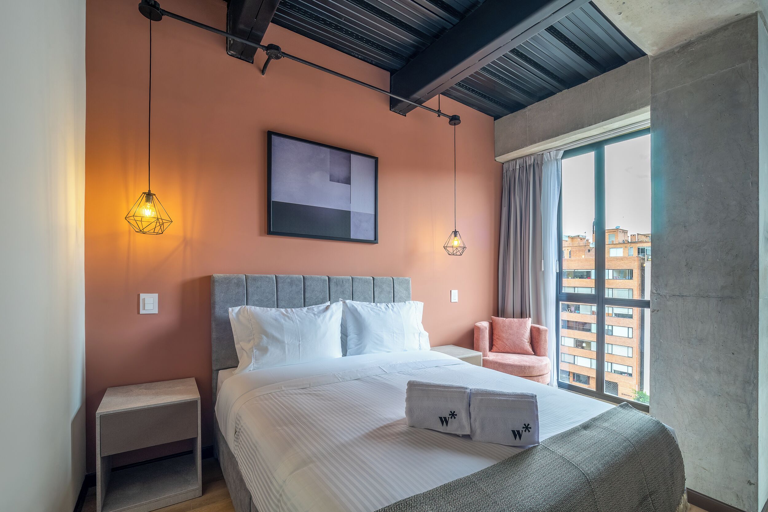 A modern hotel room with a large bed, two bedside lamps, a window with a view of buildings, and a grey headboard.