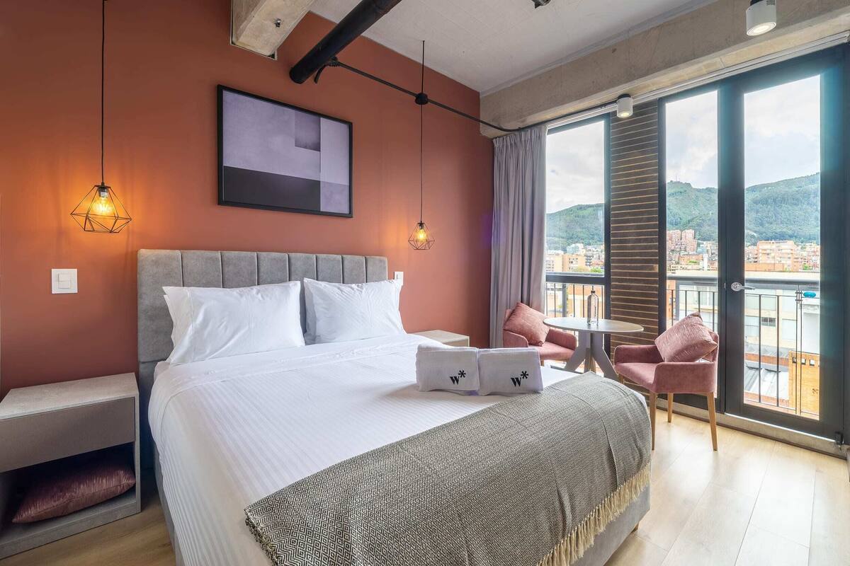 Habitación de hotel con una cama grande, un escritorio, una silla y un balcón con vista a la ciudad.