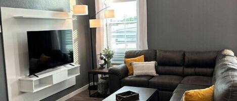 Una sala moderna con un televisor de pantalla plana montado en una pared blanca, un sofá de cuero oscuro con cojines, una mesa de centro y un tapete estampado.