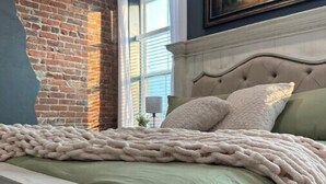 A bedroom with a brick wall, a large window with white curtains, a bed with a green bedspread, and a painting above the bed.