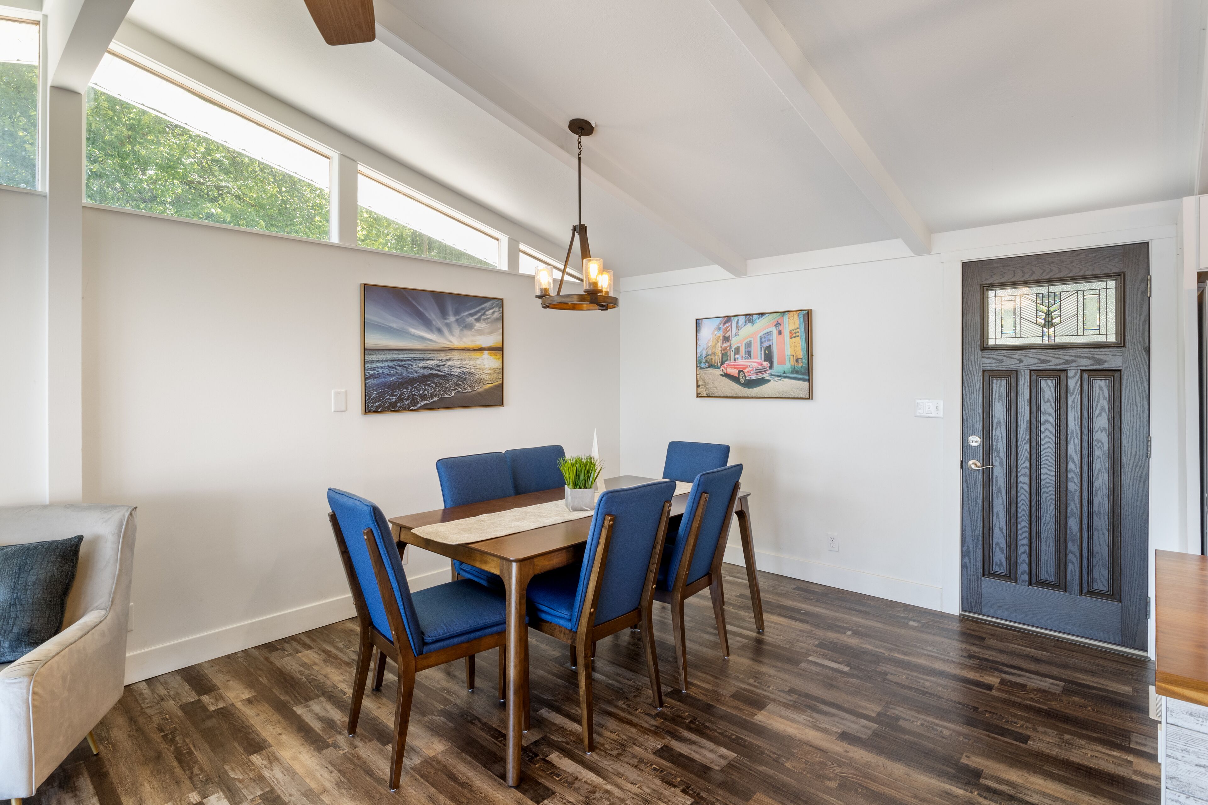 Een eetgedeelte met een houten tafel, blauwe stoelen en een kroonluchter. Op de muren hangen schilderijen en er is een deur met een raam.