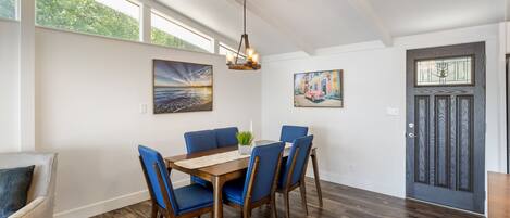 Een eetgedeelte met een houten tafel, blauwe stoelen en een kroonluchter. Op de muren hangen schilderijen en er is een deur met een raam.