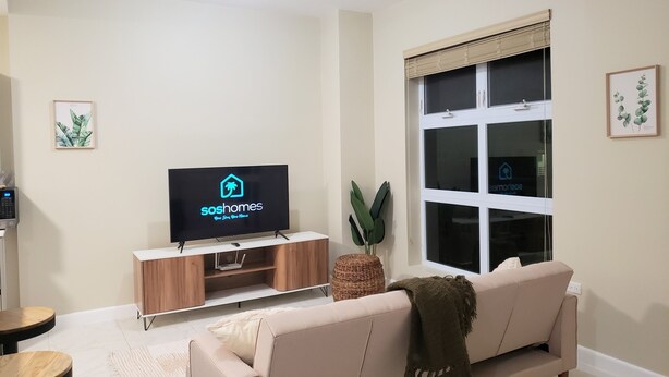 A living room with a TV displaying 'soshomes' logo, a sofa, a small table, and a window with blinds. - Lofts 1B/R Lounge w/WiFi|Gym|near National Stadium (Kingston)
