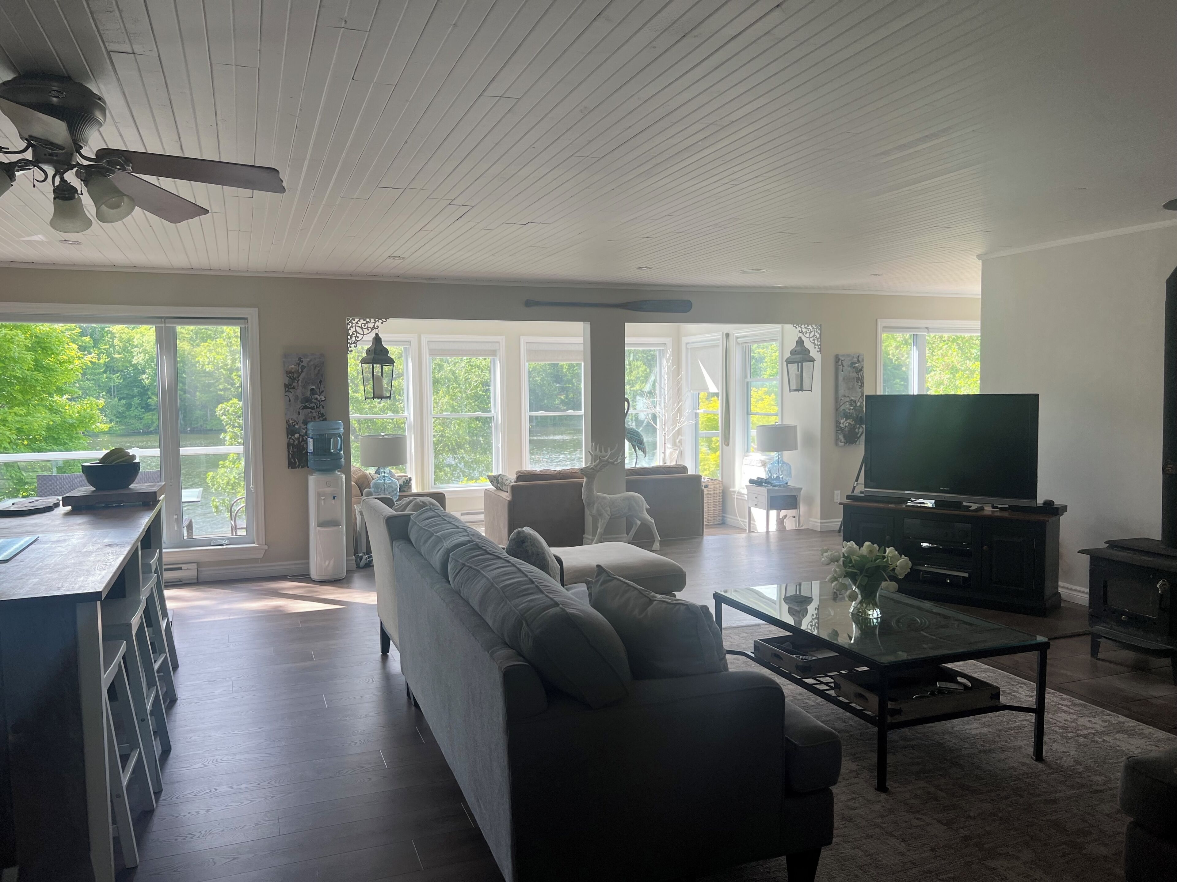 A spacious living room with a sofa, coffee table, and television. Large windows provide a view of the outdoors.