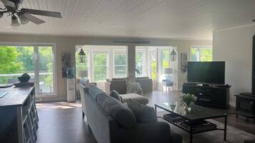 A spacious living room with a sofa, coffee table, and television. Large windows provide a view of the outdoors.