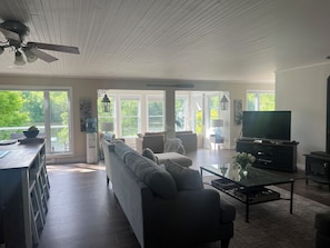A spacious living room with a sofa, coffee table, and television. Large windows provide a view of the outdoors.