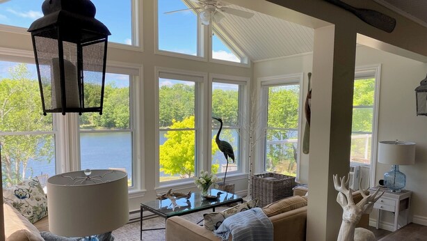 A spacious living room with large windows, a ceiling fan, a decorative lamp, a glass coffee table, and a plush sofa. - Luxury cottage on beautiful lakefront with hot-tub & private bunkie. New to VRBO (Maberley)