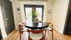 Un coin repas avec une table ronde, quatre chaises en bois, un vase avec des fleurs et une vue sur une piscine par la fenêtre.