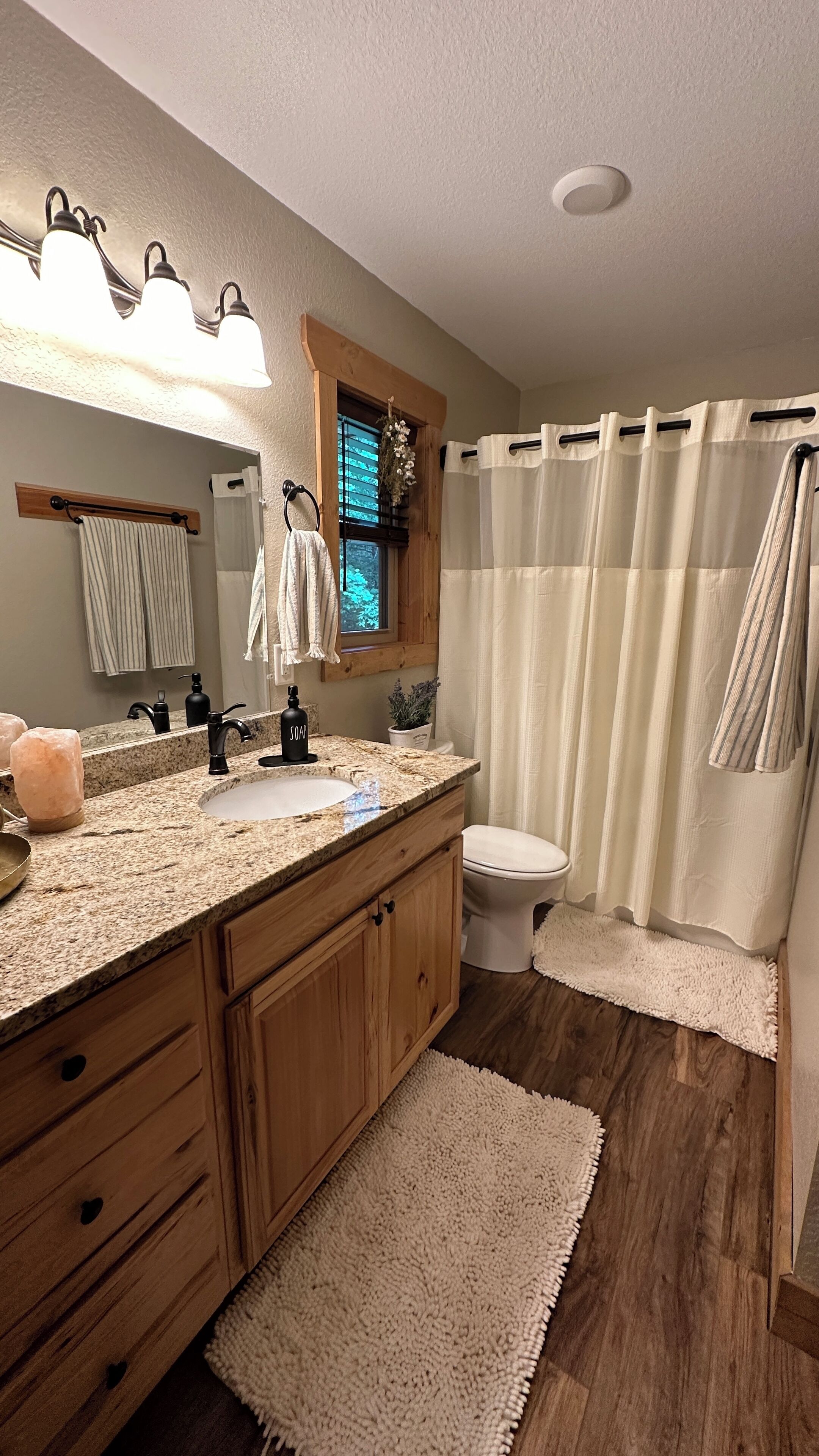 A bathroom with a granite countertop sink, wooden cabinets, a mirror, a window with blinds, a toilet, and a shower with a curtain.