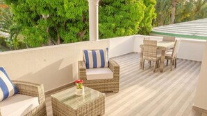Un balcón con mobiliario de mimbre, incluyendo un sofá y sillas, una pequeña mesa con un florero y vistas a árboles verdes y frondosos.