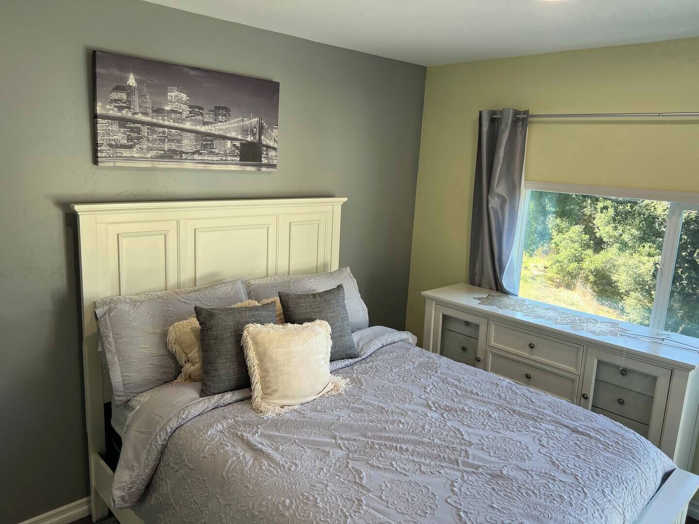 A bedroom with a bed, a nightstand, a window with curtains, and a cityscape painting on the wall.