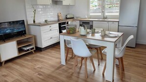 Une cuisine moderne avec des meubles blancs, une table à manger avec des chaises, un téléviseur posé sur une étagère et un réfrigérateur.