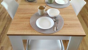 Une table à manger est dressée avec des assiettes, des bols et des couverts, entourée de chaises blanches dans une cuisine moderne. On aperçoit un téléviseur et des placards au fond de la pièce.