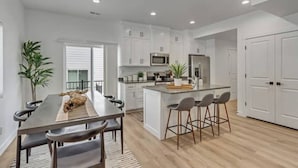 Una cocina moderna con armarios blancos, una isla central con taburetes y un comedor con una mesa larga y sillas.