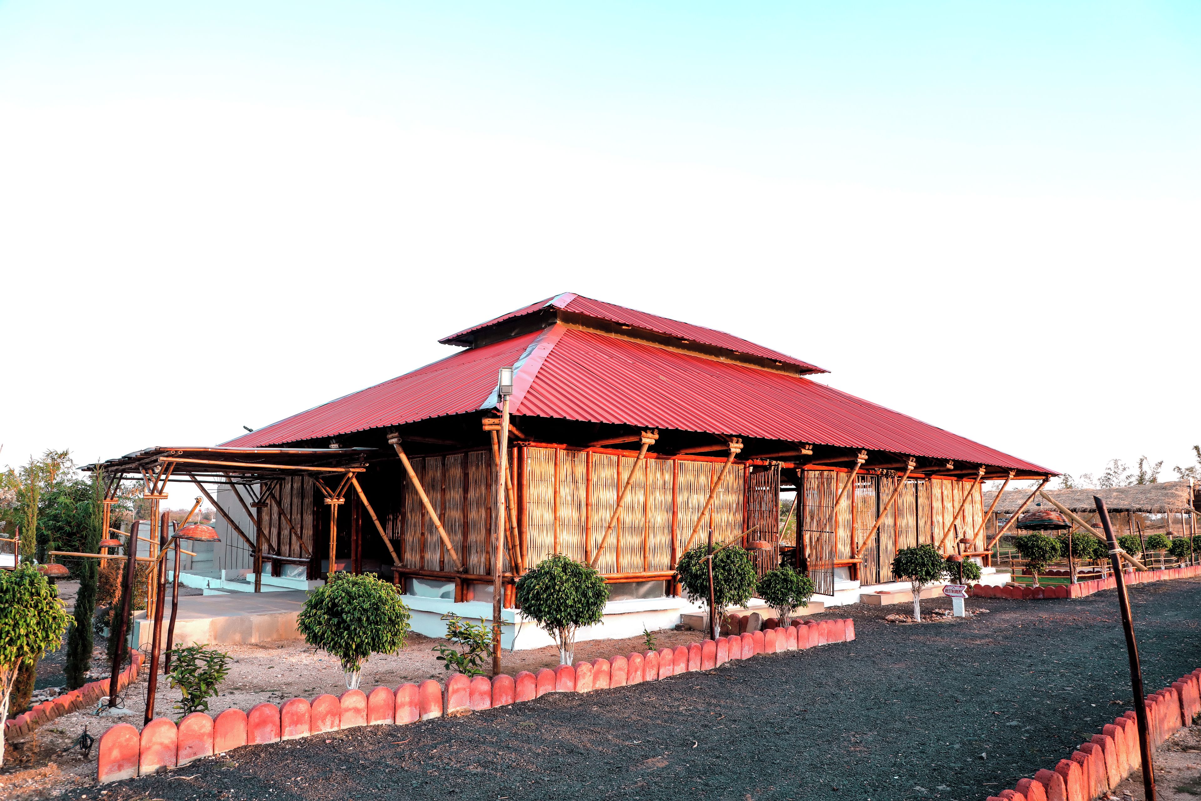 A building with a red roof and bamboo walls, surrounded by a red fence and small trees.