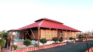 A building with a red roof and bamboo walls, surrounded by a red fence and small trees.