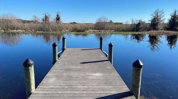 A wooden pier extending into calm water with clear reflections, surrounded by trees and vegetation.