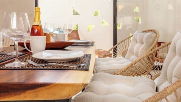 Ein Essbereich mit einem Holztisch, gedeckt für zwei Personen, inklusive Weingläsern, einer Flasche und Tellern. Wicker-Stühle mit Kissen sind vorhanden.