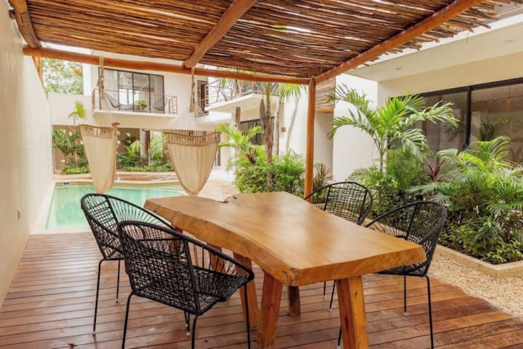 A wooden table with four chairs under a thatched roof, surrounded by lush greenery and a pool.